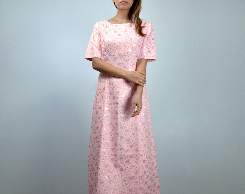 May include: A light pink, floor-length dress with short sleeves. The dress features a subtle floral pattern in a lighter shade of pink and silver. The dress has a boat neckline and a simple, elegant design.