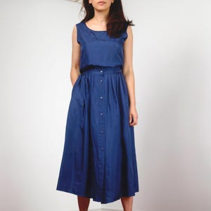 May include: A sleeveless, navy blue dress with a button-down skirt. The dress features a round neckline and a fitted bodice that flows into a pleated skirt. The model is wearing the dress in front of a white background.