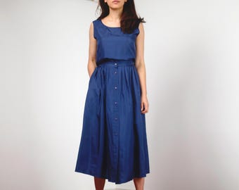 May include: A sleeveless, navy blue dress with a button-down skirt. The dress features a round neckline and a fitted bodice that flows into a pleated skirt. The model is wearing the dress in front of a white background.