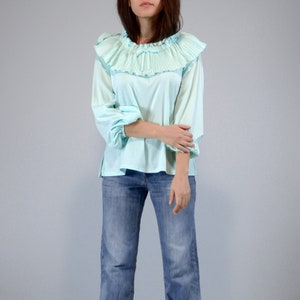 Pode incluir: Uma blusa azul clara com gola franzida e mangas compridas. A blusa &eacute; combinada com jeans azuis. A gola franzida adiciona um toque vintage &agrave; parte de cima. As mangas s&atilde;o franzidas nos punhos.