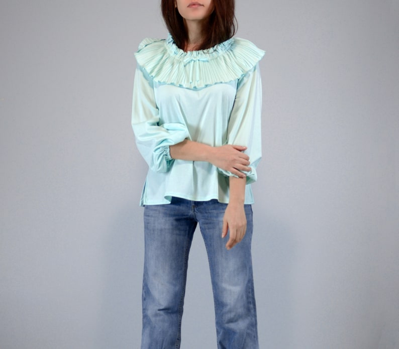 Pode incluir: Uma blusa azul clara com gola franzida e mangas compridas. A blusa &eacute; combinada com jeans azuis. A gola franzida adiciona um toque vintage &agrave; parte de cima. As mangas s&atilde;o franzidas nos punhos.