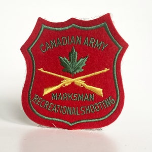 May include: Red felt patch with a green embroidered border. The patch features a green maple leaf, two crossed gold rifles, and the text "Canadian Army Marksman Recreational Shooting".