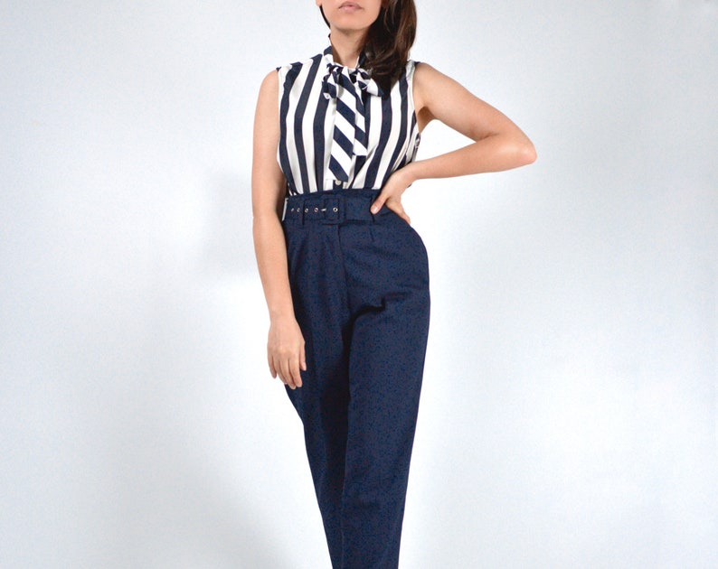 May include: A woman wearing a white and black striped sleeveless blouse with a bow tie and navy blue high-waisted pants with a belt.