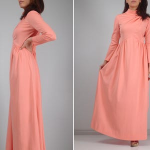 K&ouml;nnte beinhalten: Ein langes, pfirsichfarbenes Kleid mit langen &Auml;rmeln und hohem Ausschnitt. Das Kleid hat ein eng anliegendes Oberteil und einen flie&szlig;enden Rock. Der Stoff scheint leicht zu sein. Das Kleid ist einfarbig und hat einen Vintage-Stil.