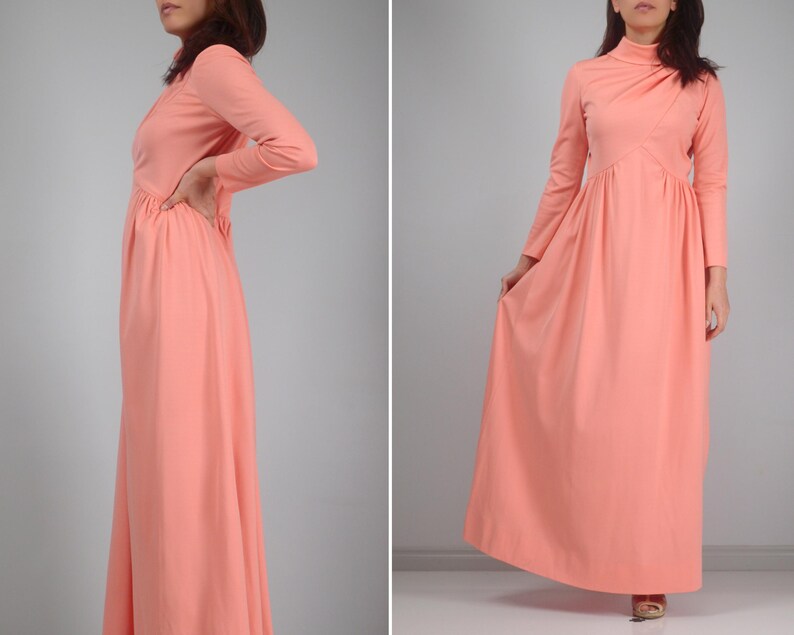 K&ouml;nnte beinhalten: Ein langes, pfirsichfarbenes Kleid mit langen &Auml;rmeln und hohem Ausschnitt. Das Kleid hat ein eng anliegendes Oberteil und einen flie&szlig;enden Rock. Der Stoff scheint leicht zu sein. Das Kleid ist einfarbig und hat einen Vintage-Stil.