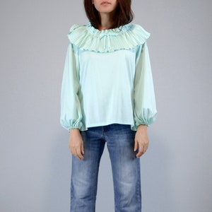 Pode incluir: Uma blusa azul clara com gola de babados plissados e mangas compridas. As mangas s&atilde;o franzidas nos pulsos. Combinada com cal&ccedil;as de ganga azuis. O fundo &eacute; cinza claro liso.