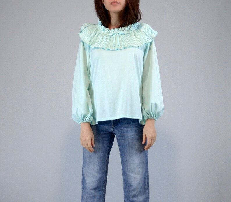 Pode incluir: Uma blusa azul clara com gola de babados plissados e mangas compridas. As mangas s&atilde;o franzidas nos pulsos. Combinada com cal&ccedil;as de ganga azuis. O fundo &eacute; cinza claro liso.