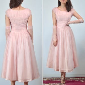 May include: A light pink vintage dress with a fitted bodice and a full, flowing skirt. The dress features short sleeves and matching sheer gloves. The fabric appears to be a delicate tulle, creating a soft, romantic look. The dress is knee-length.