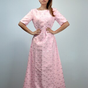 May include: A light pink, floor-length dress with short sleeves and a boat neckline. The dress features a subtle floral pattern and a fitted bodice that flows into a flared skirt. The fabric has a slight sheen, giving it a vintage look.