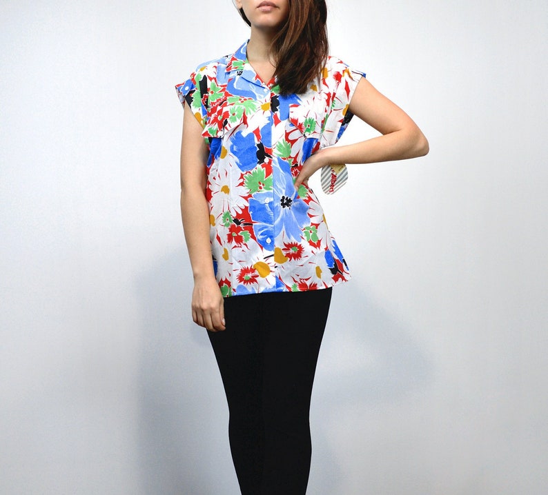 May include: A short-sleeved, button-down shirt with a vibrant floral pattern. The shirt features a white background with large blue, red, and yellow flowers. The collar and sleeves are trimmed in white. The model is wearing black pants.