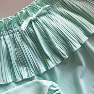 Pode incluir: Close-up de uma saia turquesa clara com um detalhe de babados plissados. A saia tem uma cintura com cord&atilde;o e um tecido macio e fluido. O design geral &eacute; simples e elegante, adequado para roupas casuais.