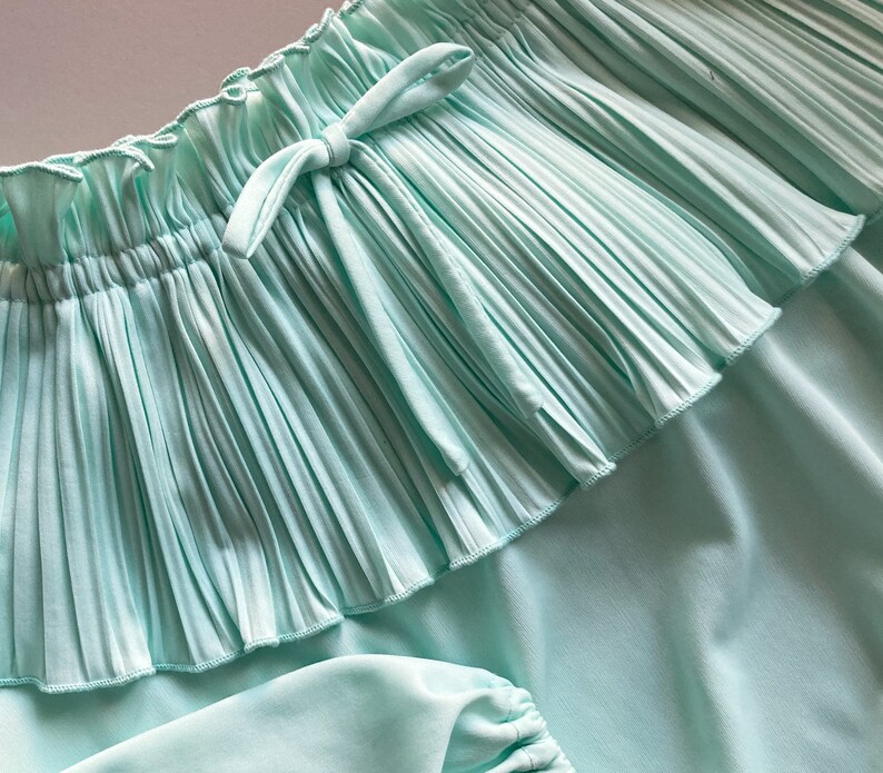 Pode incluir: Close-up de uma saia turquesa clara com um detalhe de babados plissados. A saia tem uma cintura com cord&atilde;o e um tecido macio e fluido. O design geral &eacute; simples e elegante, adequado para roupas casuais.