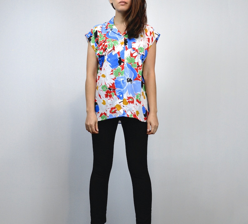 May include: A sleeveless button-down shirt with a vibrant floral pattern. The shirt features large blue, red, and yellow flowers on a white background. The model is wearing black leggings.