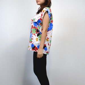 May include: A sleeveless top with a vibrant floral pattern. The top features large white daisy-like flowers with yellow centers, accented by red, blue, green, and black details. The garment is paired with black leggings, creating a striking contrast.