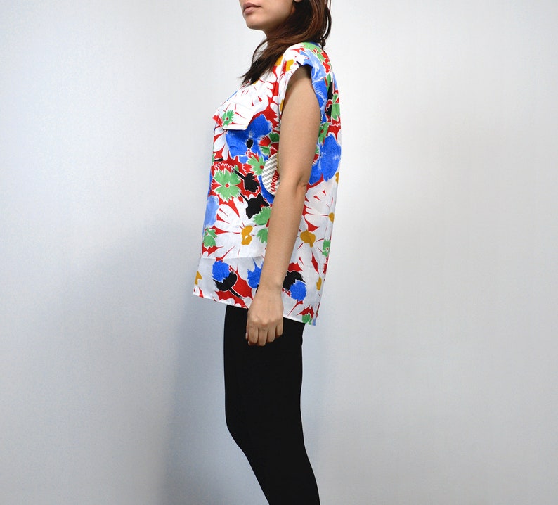 May include: A sleeveless top with a vibrant floral pattern. The top features large white daisy-like flowers with yellow centers, accented by red, blue, green, and black details. The garment is paired with black leggings, creating a striking contrast.