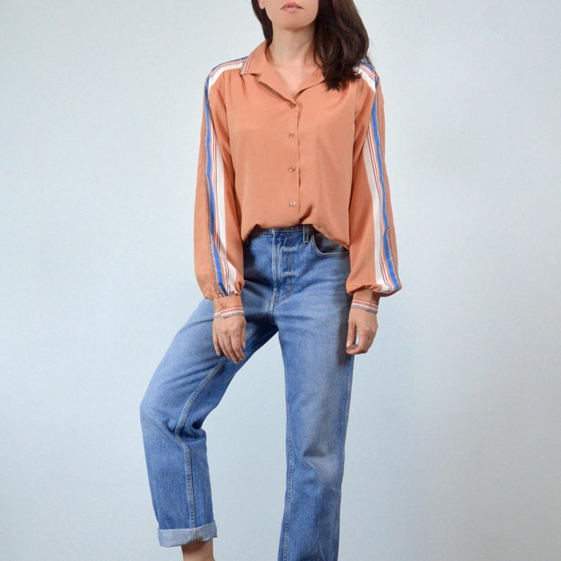 May include: A peach-colored button-down shirt with long sleeves and blue and white stripes down the arms. The shirt is paired with light blue denim jeans.