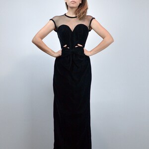 May include: A black velvet evening gown with a sheer neckline and cap sleeves. The bodice features a sweetheart neckline and cut-out details at the waist, with a bow detail. The gown has a floor-length skirt.