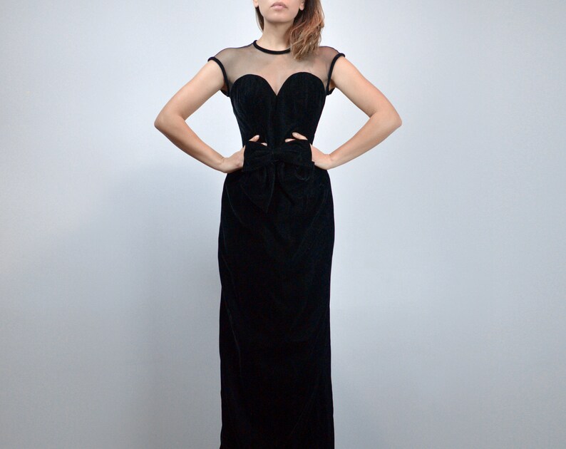 May include: A black velvet evening gown with a sheer neckline and cap sleeves. The bodice features a sweetheart neckline and cut-out details at the waist, with a bow detail. The gown has a floor-length skirt.