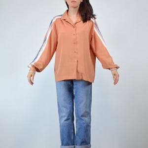 May include: A peach-colored button-down shirt with white and blue stripes on the sleeves. The shirt is paired with blue jeans. The sleeves have a cuff detail with matching stripes. The shirt has a classic collar.