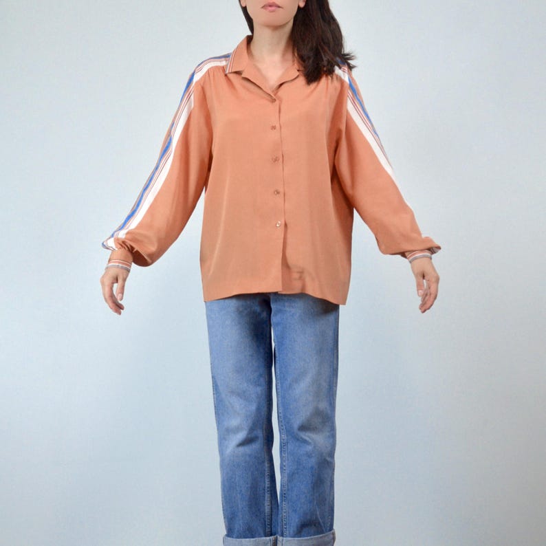 May include: A peach-colored button-down shirt with white and blue stripes on the sleeves. The shirt is paired with blue jeans. The sleeves have a cuff detail with matching stripes. The shirt has a classic collar.
