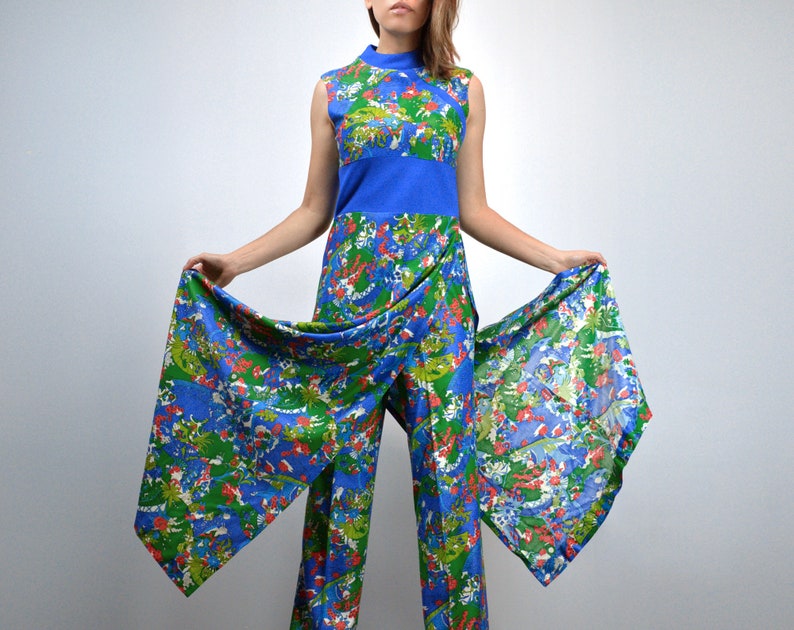 May include: A sleeveless jumpsuit in a vibrant blue, green, and red floral pattern. The jumpsuit features a high neckline, a blue waist panel, and wide-leg trousers with attached fabric panels that extend outward. The fabric has a vintage aesthetic.