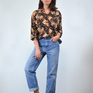 May include: A black and gold patterned long-sleeved button-up shirt with a collared neckline. The shirt is worn with light blue jeans.
