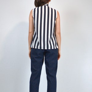 May include: A sleeveless striped blouse with a white background and black stripes. The blouse has a high neckline and a relaxed fit. The model is wearing dark blue pants.