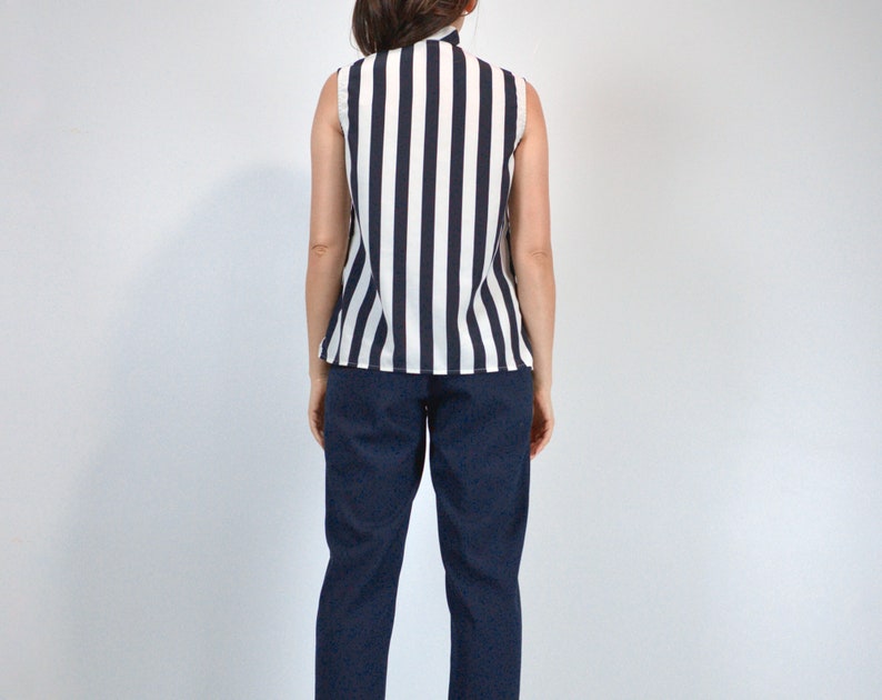 May include: A sleeveless striped blouse with a white background and black stripes. The blouse has a high neckline and a relaxed fit. The model is wearing dark blue pants.