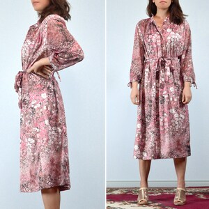 May include: A vintage midi dress in a floral print. The dress features a button-down front, long sleeves with tie details at the cuffs, and a matching waist tie. The fabric has a pink, white, and black floral pattern.