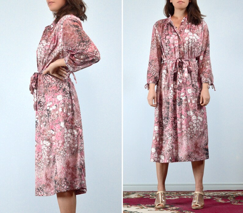 May include: A vintage midi dress in a floral print. The dress features a button-down front, long sleeves with tie details at the cuffs, and a matching waist tie. The fabric has a pink, white, and black floral pattern.