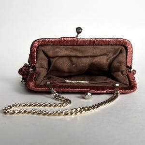 May include: Open, vintage-style purse in a reddish-brown hue, featuring a gold chain strap. The interior is a darker brown fabric. The purse has a metal clasp and a decorative element on the side.