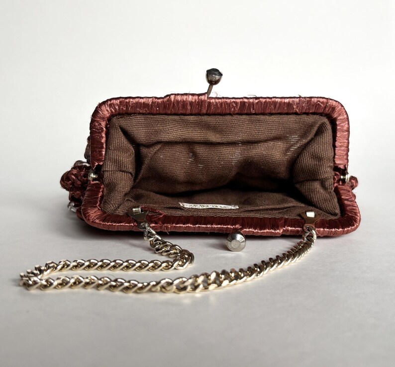 May include: Open, vintage-style purse in a reddish-brown hue, featuring a gold chain strap. The interior is a darker brown fabric. The purse has a metal clasp and a decorative element on the side.