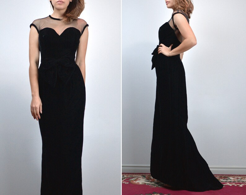 May include: A black velvet evening gown with a sweetheart neckline and sheer cap sleeves. The dress features a fitted bodice, a cinched waist with a bow detail, and a floor-length skirt. The gown is a classic, elegant design.