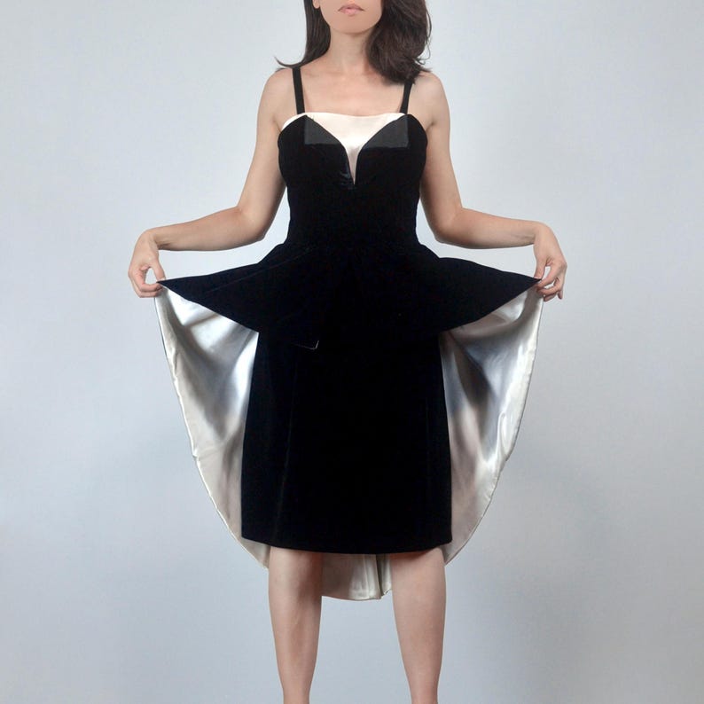 May include: A black velvet dress featuring a sweetheart neckline with a white satin insert, thick shoulder straps, and a peplum waist. The skirt has a high-low hemline, revealing a smooth, reflective white satin lining underneath the black velvet.