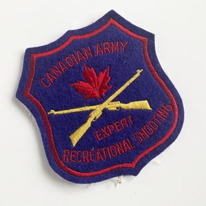 May include: A blue felt patch with red and yellow embroidery. It reads "Canadian Army Expert Recreational Shooting". A red maple leaf and two crossed rifles are also embroidered on the patch.