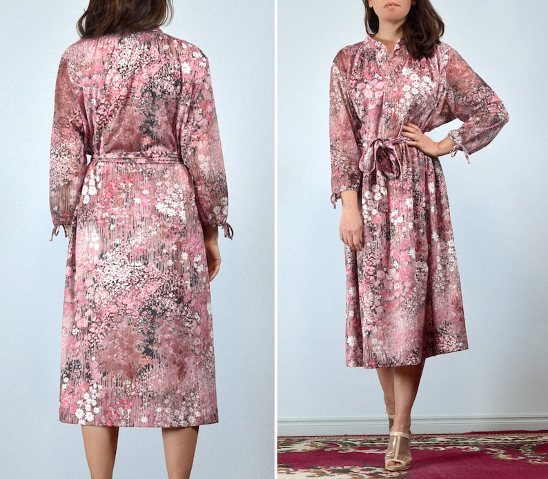 May include: A vintage midi dress featuring a floral pattern in shades of pink, white, and black. The dress has long sleeves with tie details at the cuffs, a tie waist, and a collared neckline. The dress is knee-length.