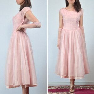 May include: A light pink vintage dress with a fitted bodice and a full, flowing skirt. The dress features short sleeves and is made of a sheer, layered fabric. The dress is knee-length and has a delicate, romantic style.