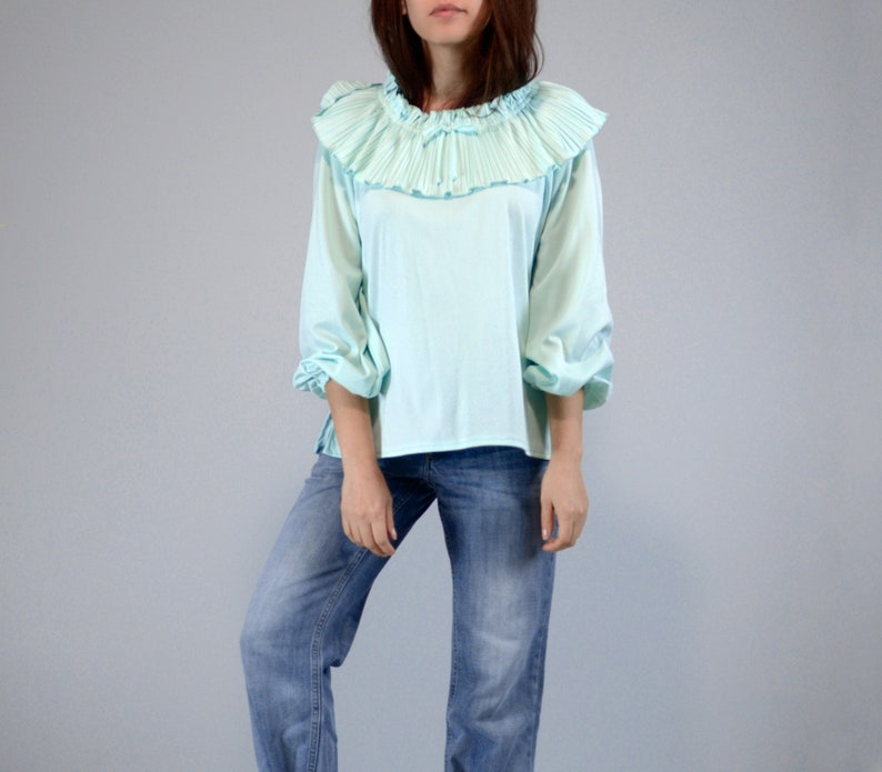 Pode incluir: Blusa azul clara com gola franzida e mangas tr&ecirc;s quartos. A blusa tem um corte solto e &eacute; combinada com jeans azuis. O fundo &eacute; cinza claro liso.