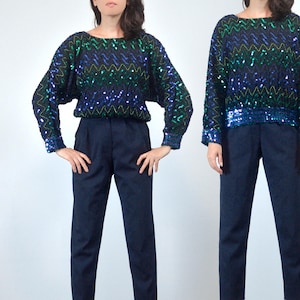 May include: A sequinned top with a zig-zag pattern in shades of green, blue, and black, paired with navy blue trousers. The top has long sleeves and a round neckline. The trousers are high-waisted with a straight leg.
