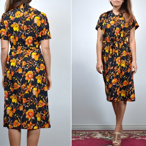 Puede incluir: Un vestido midi negro con estampado floral en tonos naranja, amarillo y marr&oacute;n. El vestido tiene manga corta y un lazo en la cintura.