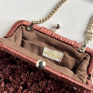 May include: Close-up of a vintage maroon handbag with a gold chain strap. The bag features a woven texture and a clasp closure. The interior is lined with brown fabric, and a tag inside reads "ARTEL MFG CO."