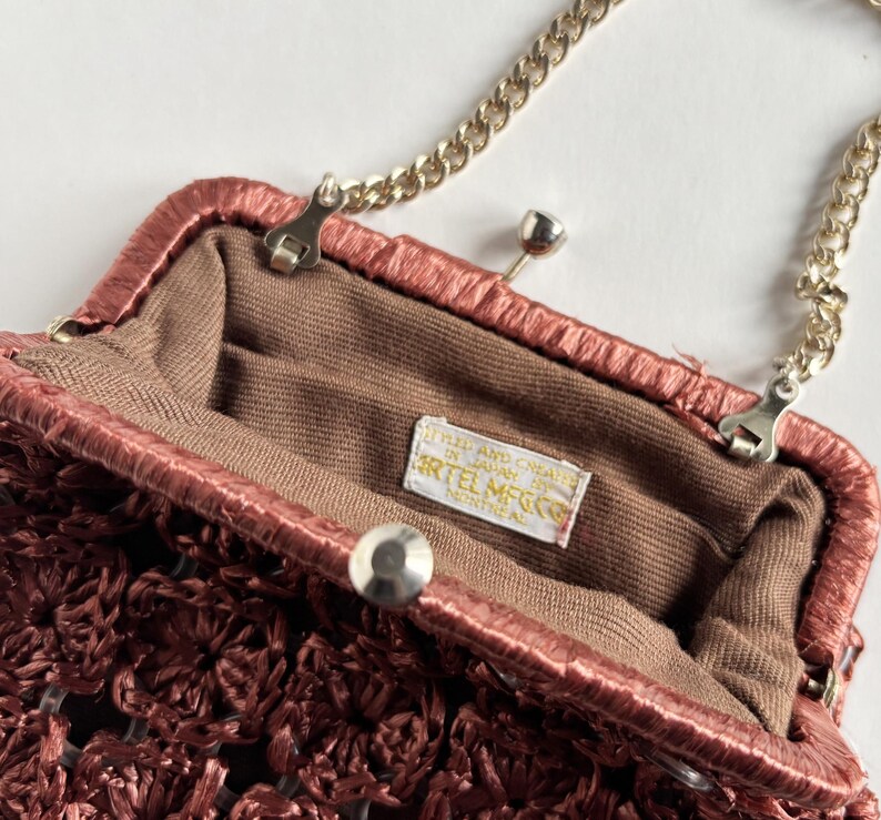 May include: Close-up of a vintage maroon handbag with a gold chain strap. The bag features a woven texture and a clasp closure. The interior is lined with brown fabric, and a tag inside reads "ARTEL MFG CO."