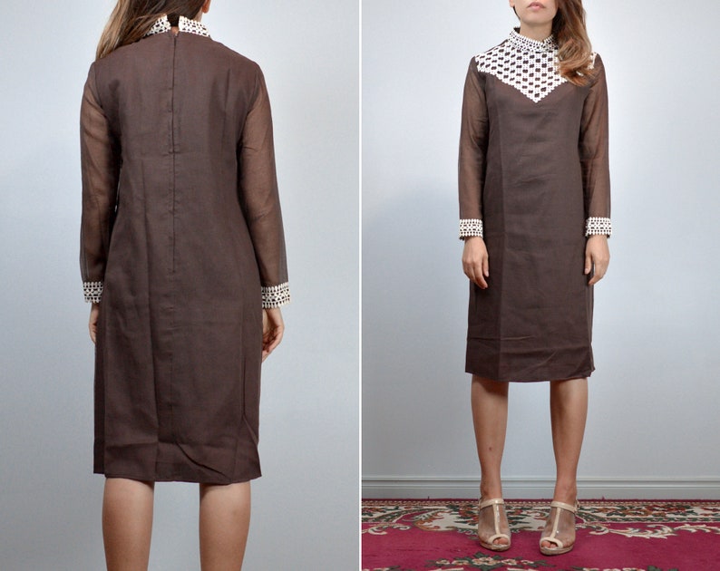 May include: A brown vintage dress with long sheer sleeves and a high collar. The collar and cuffs feature a white and brown geometric pattern. The dress has a knee-length hemline and a simple, elegant silhouette, perfect for a retro look.