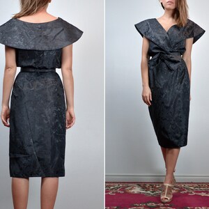 May include: A black midi dress featuring a subtle tonal floral pattern, a wide collar that extends over the shoulders, a V-neckline, and a matching fabric belt tied at the waist. The dress has a structured silhouette with short, flared sleeves.