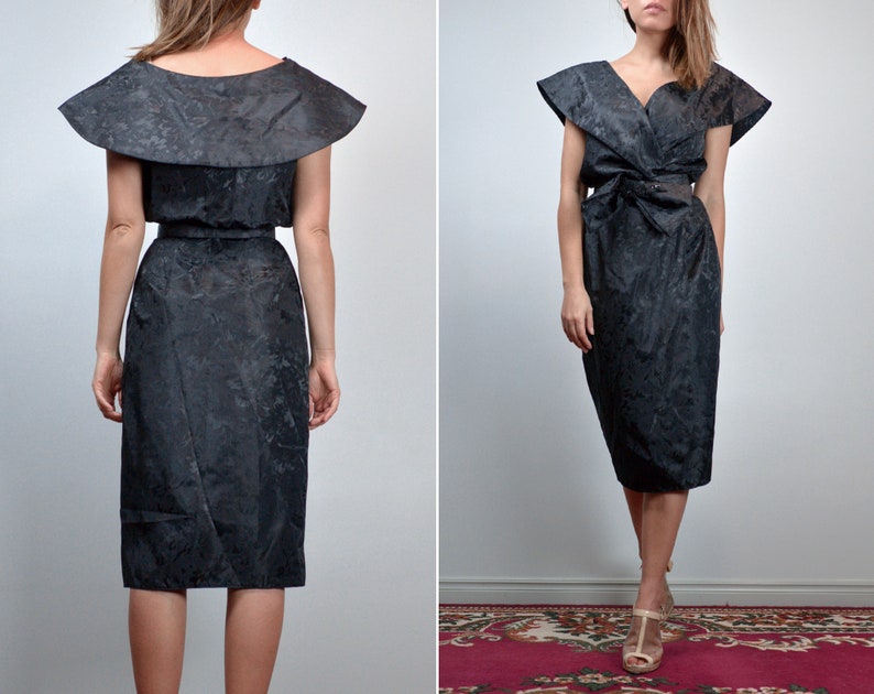 May include: A black midi dress featuring a subtle tonal floral pattern, a wide collar that extends over the shoulders, a V-neckline, and a matching fabric belt tied at the waist. The dress has a structured silhouette with short, flared sleeves.