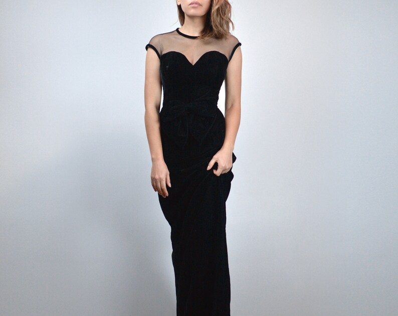 May include: A black velvet evening gown with a sheer neckline and cap sleeves. The bodice features a sweetheart neckline and a fitted waist, flowing into a floor-length skirt. The dress is a classic, elegant design.