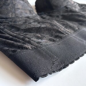 May include: Close-up of a black bra with a delicate floral lace pattern. The bra features a structured design with a wide, ribbed band at the bottom and scalloped edges. The fabric has a subtle sheen, and the overall aesthetic is elegant and refined.
