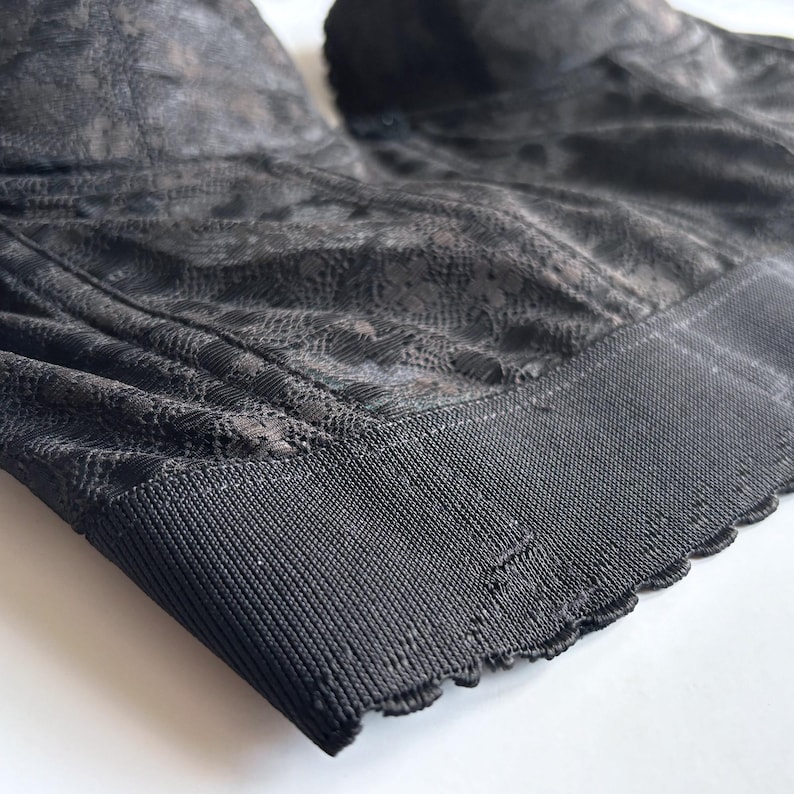 May include: Close-up of a black bra with a delicate floral lace pattern. The bra features a structured design with a wide, ribbed band at the bottom and scalloped edges. The fabric has a subtle sheen, and the overall aesthetic is elegant and refined.
