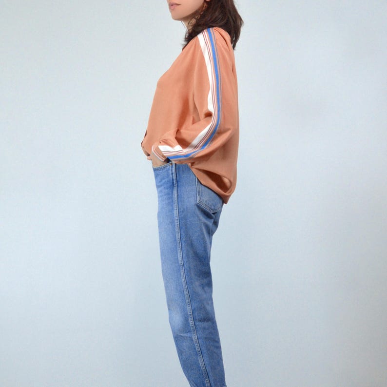 May include: A light orange jacket with white and blue stripes down the sleeves. The jacket is paired with blue jeans. The outfit is casual and stylish.