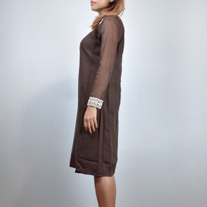 May include: A brown dress with long, sheer sleeves and a knee-length hemline. The dress features a white, textured cuff detail on the sleeves. The dress is made of a lightweight fabric and has a simple, elegant design.
